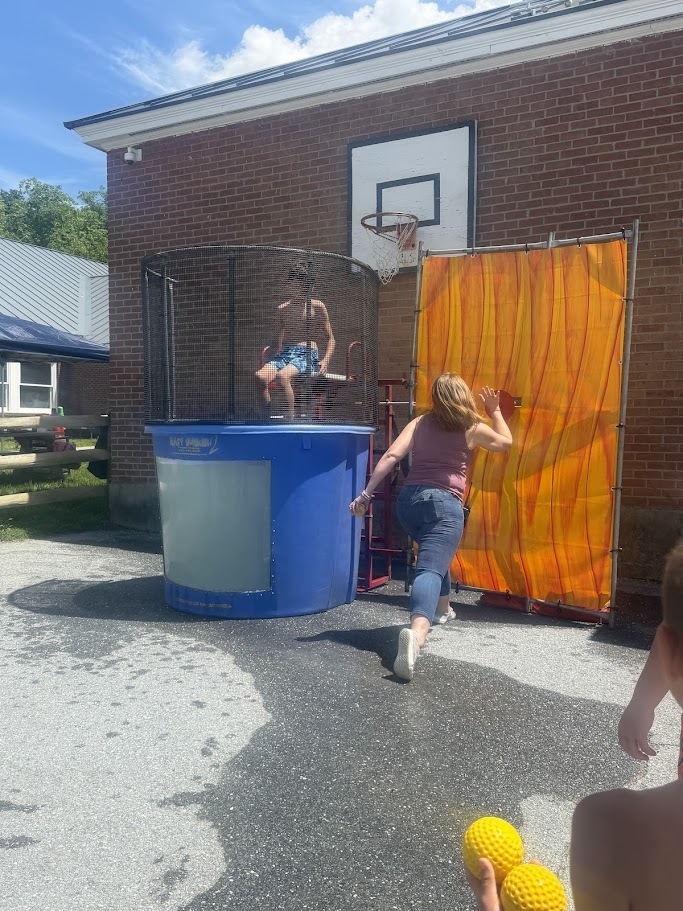 A staff member throws a ball at a dunk tank target while a student sits above the tank waiting to see if the throw will send them into the water. Photo 4