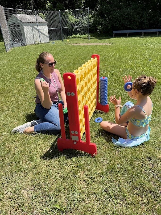 A staff member and student sit on the grass playing a giant outdoor Connect Four game during a school field day.