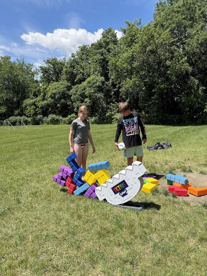 Two students play with oversized Tetris-style blocks on the grass, stacking colorful pieces during an outdoor field day activity.