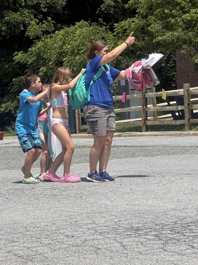 A staff member leads two students across the field during an outdoor activity, pointing toward the next station while carrying supplies and towels.