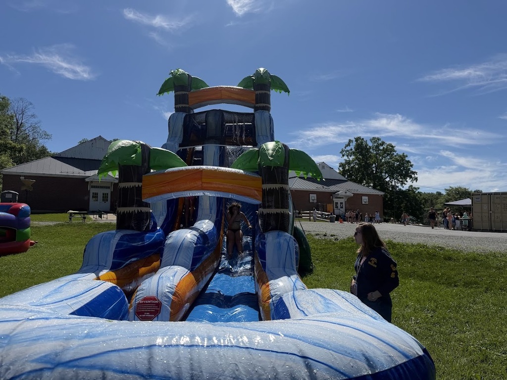 A student exits a large tropical-themed inflatable water slide during a school field day while a staff member stands nearby supervising. Other activities are visible in the background on a sunny day.