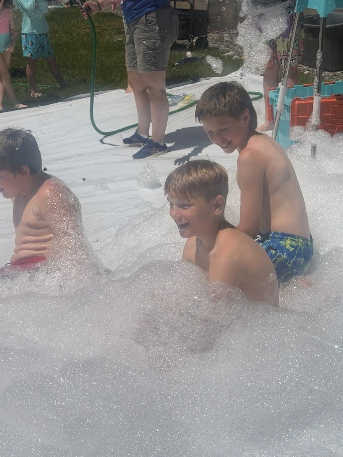 Three students sit and laugh in a large foam-filled area as bubbles surround them during a summer field day celebration.