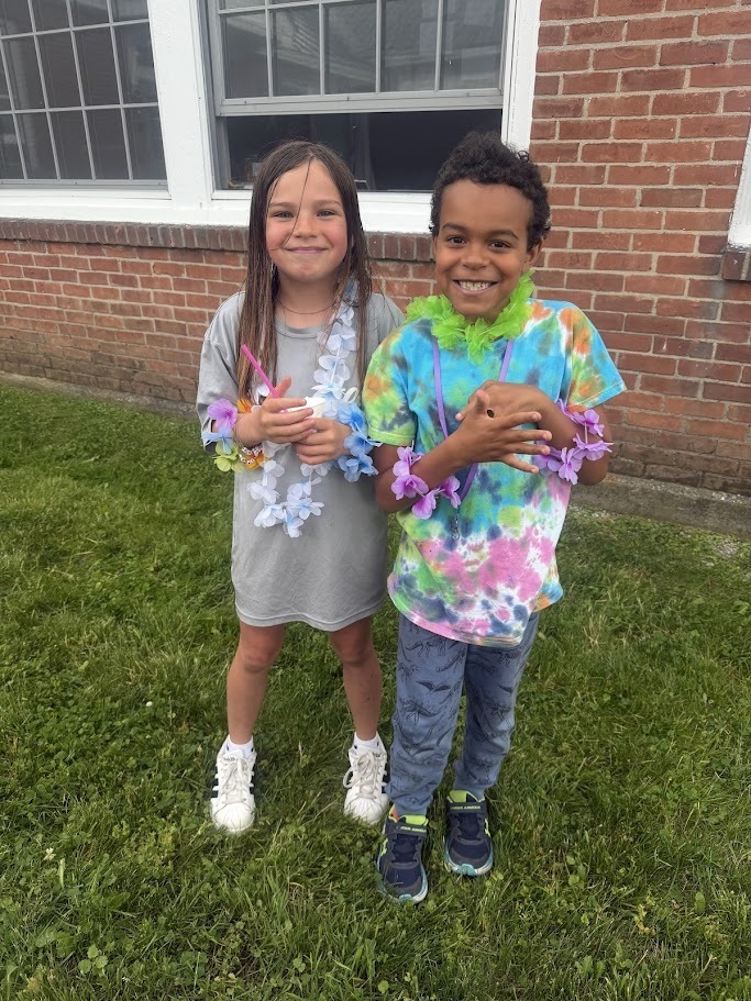 Two students stand side by side on the grass wearing colorful leis and summer-themed clothing, smiling for a photo outside the school building.