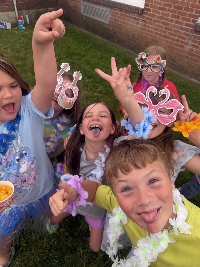 Several students wearing colorful leis and novelty sunglasses crowd around the camera, laughing, making silly faces, and showing off their festive accessories.