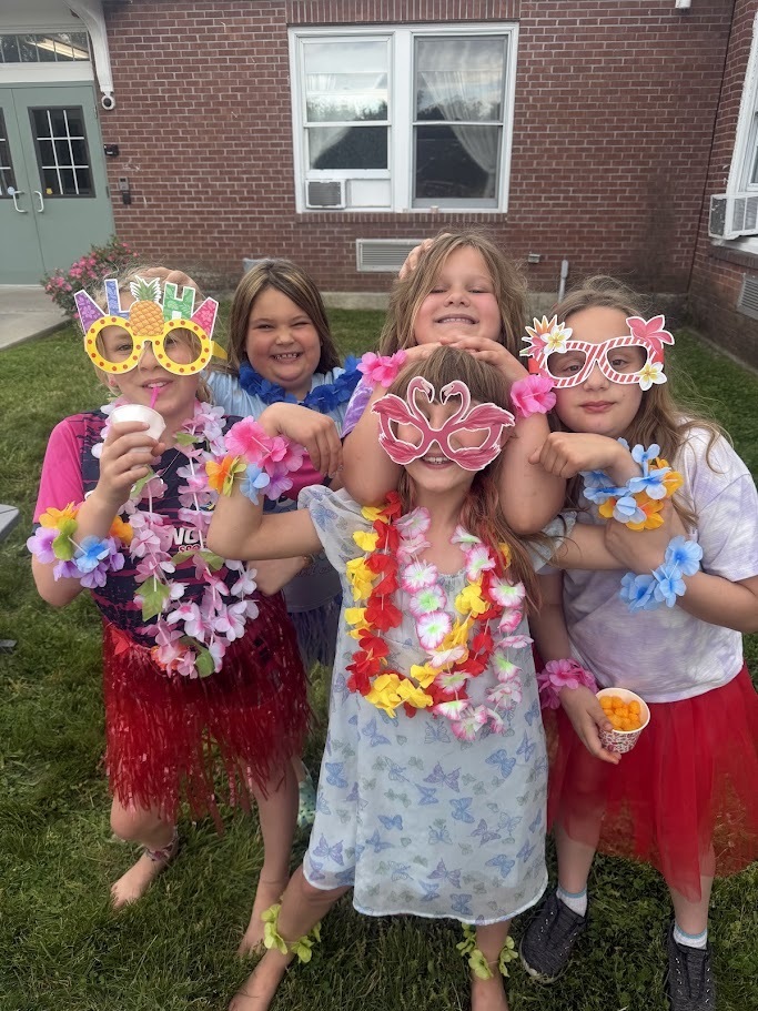 Five students dressed in Hawaiian-themed attire and colorful leis gather together outdoors, smiling and posing for a photo during a school celebration.
