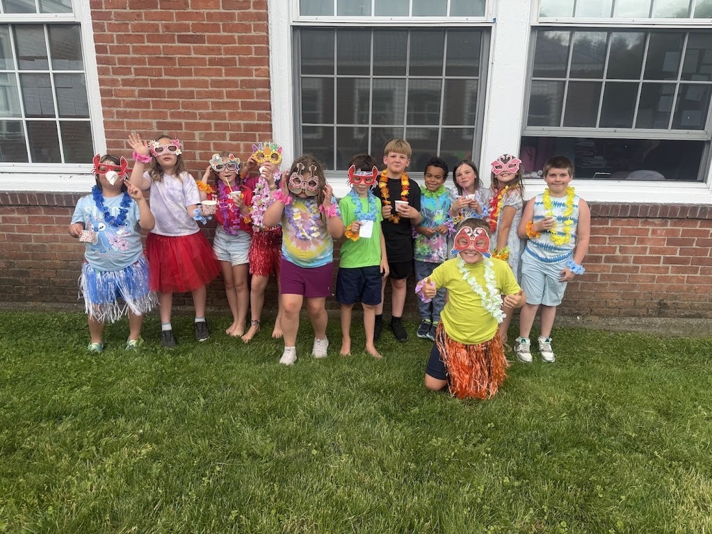 A group of elementary students pose outside in colorful Hawaiian-themed outfits, leis, grass skirts, and novelty sunglasses during a festive end-of-year celebration.