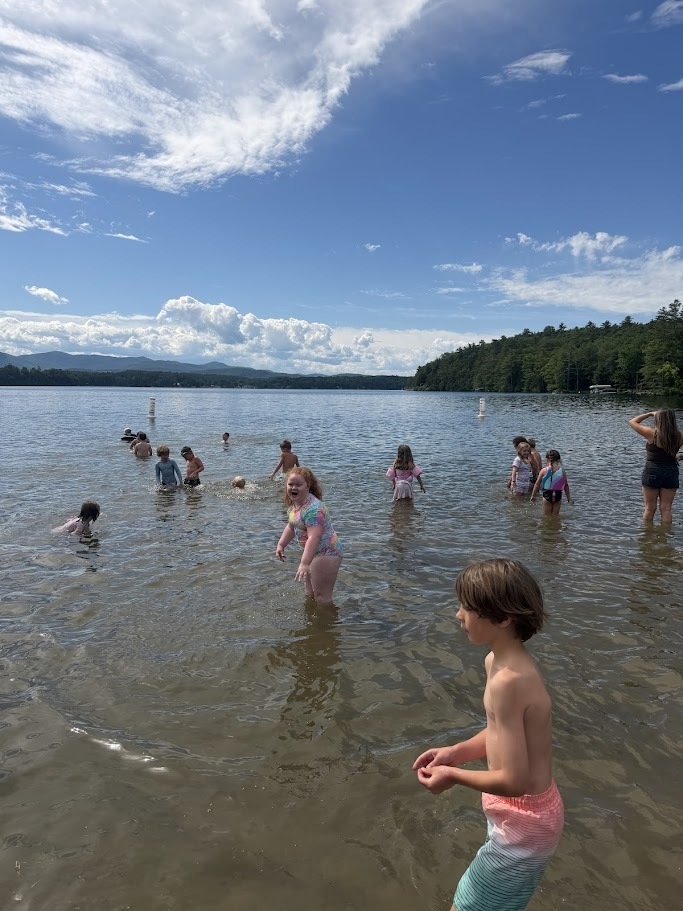 Children spread out in the shallow waters of a lake, laughing and splashing while enjoying a warm day outdoors with scenic mountains in the background.