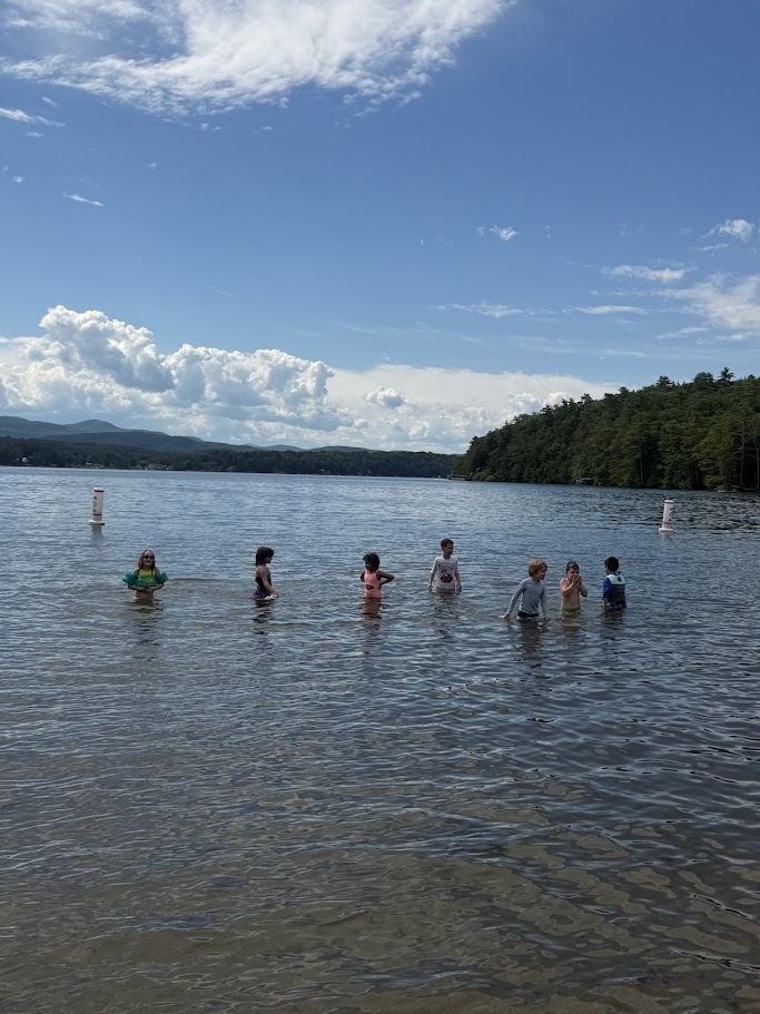 Several elementary students stand and play in the shallow water of a lake on a sunny day, with forested shorelines and mountains visible in the distance.