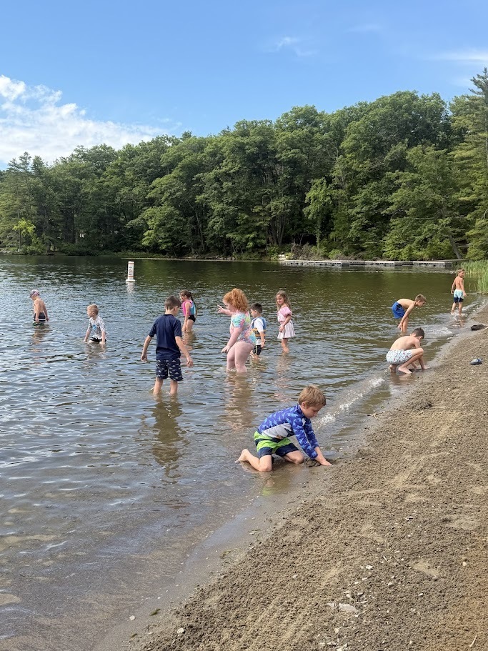 Students explore the shoreline of a lake, wading in the water, digging in the sand, and splashing near the beach during a summer field trip.