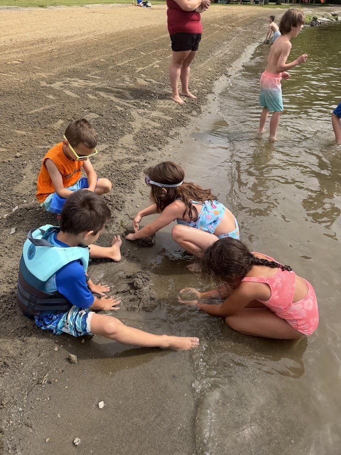 Four young students kneel at the water’s edge, digging and building with wet sand while classmates play nearby along the lakeshore.