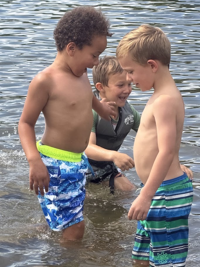 Three students laugh and play in knee-deep lake water, smiling as they splash and interact with one another near the shoreline.