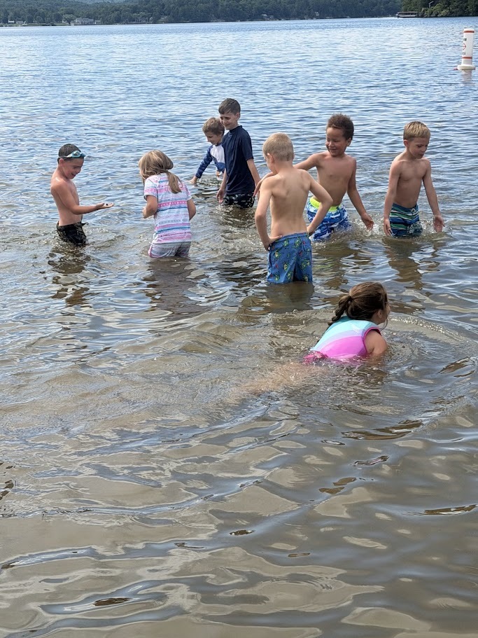 A group of children wade through shallow lake water, smiling, splashing, and exploring together during a day at the beach.