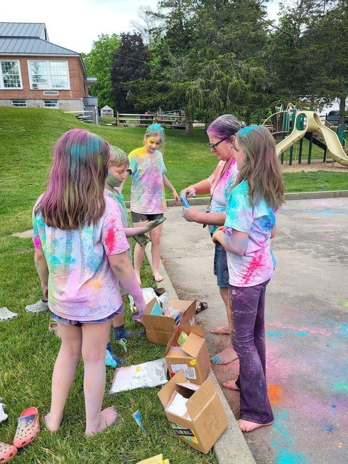 A group of students covered in colorful powder gathers around boxes of powder supplies while preparing for the next round of an outdoor color celebration.