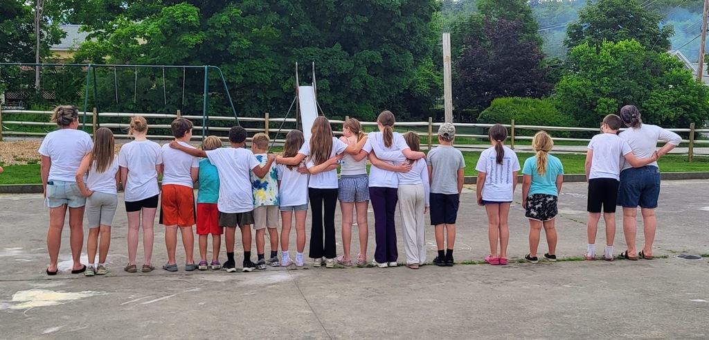 Students and teachers stand with their backs to the camera, arms around one another on the playground, posing for a group photo before an outdoor celebration.