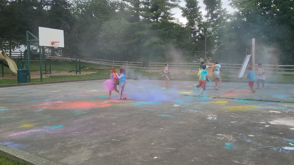 Students run and play on a playground covered in bright colored powder, creating colorful clouds as part of an outdoor end-of-year celebration.