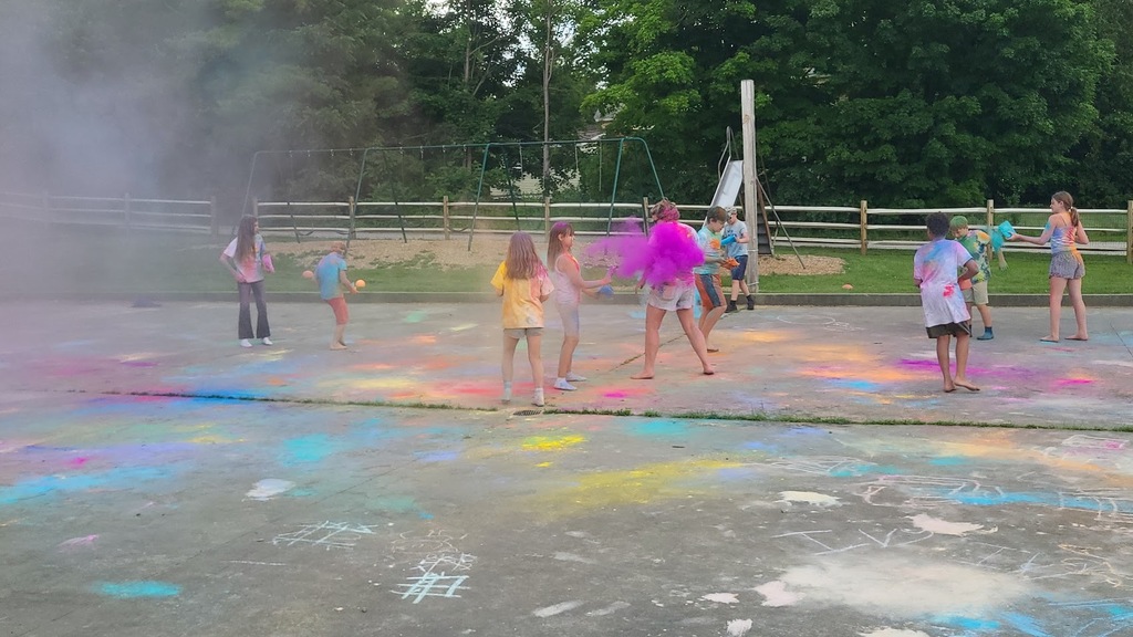 Students laugh and throw handfuls of brightly colored powder during a color run-style activity, surrounded by colorful clouds and chalk-covered pavement.