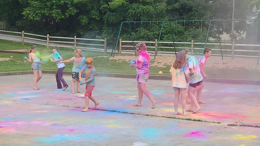 Students laugh and throw handfuls of brightly colored powder during a color run-style activity, surrounded by colorful clouds and chalk-covered pavement.