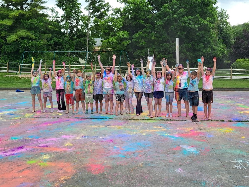 Students and staff covered head to toe in bright colored powder stand together with their hands raised, celebrating at the conclusion of a colorful outdoor event.