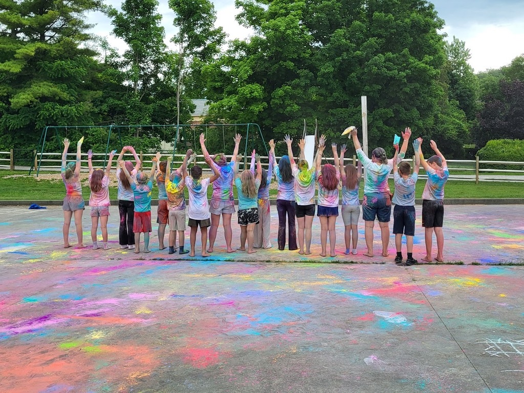 Students and staff pose with their backs to the camera and hands raised, showing shirts covered in colorful powder after an end-of-year color celebration.