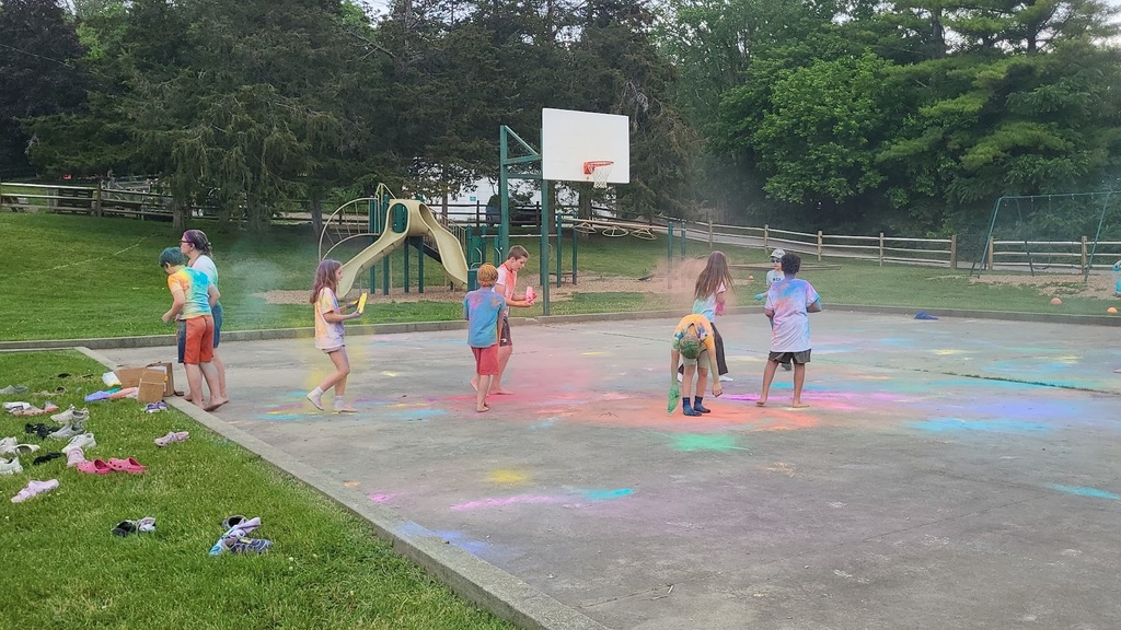 Children toss and scatter colorful powder across a paved playground, leaving vibrant splashes of pink, blue, yellow, and green on the ground.