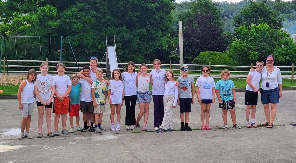 A class of elementary students and two adults stand together on a playground, smiling for a group photo while wearing white shirts before a colorful end-of-year activity.