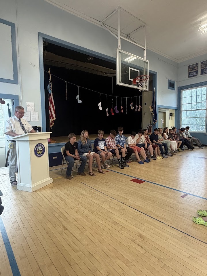 An administrator speaks at a podium during a sixth-grade recognition ceremony while students sit in a row of chairs across the front of a school gymnasium.