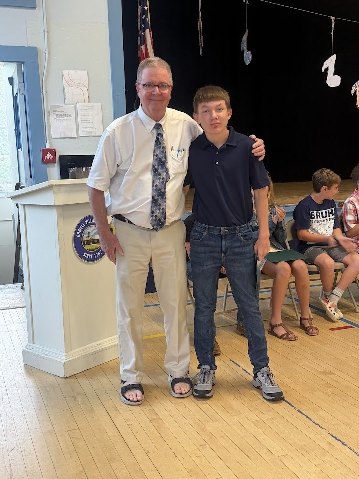 An administrator poses with a sixth-grade student in a navy polo shirt during a certificate presentation at an end-of-year awards ceremony.