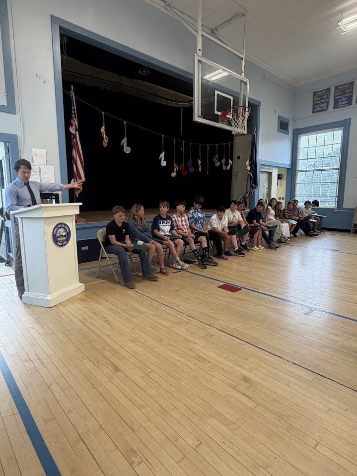 A staff member addresses students and families from a podium while sixth-grade students sit on stage-side chairs during a promotion and recognition ceremony in the school gymnasium.