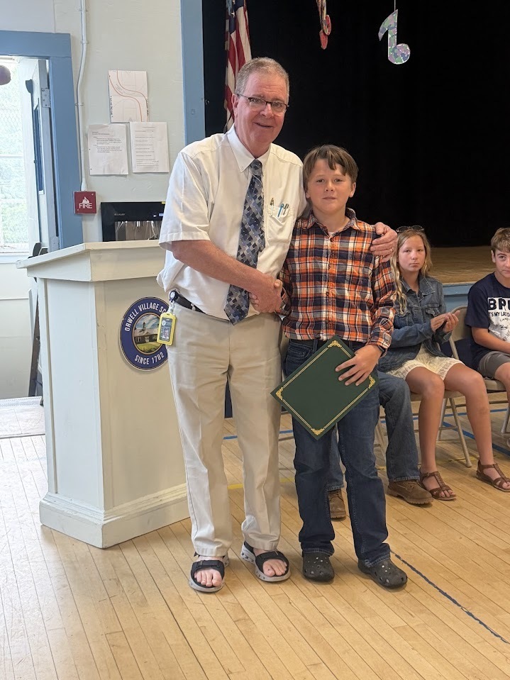 An administrator shakes hands with a sixth-grade student holding a certificate during an awards presentation at an end-of-year ceremony.