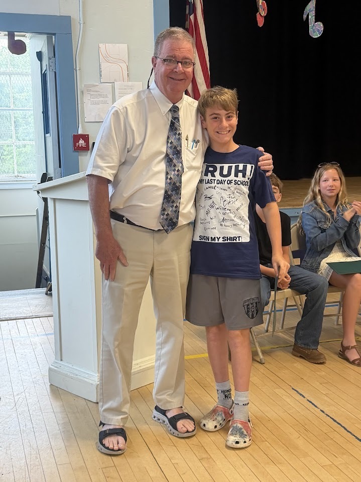 A smiling sixth-grade student wearing a signed last-day-of-school shirt poses with an administrator after receiving recognition during a school ceremony.
