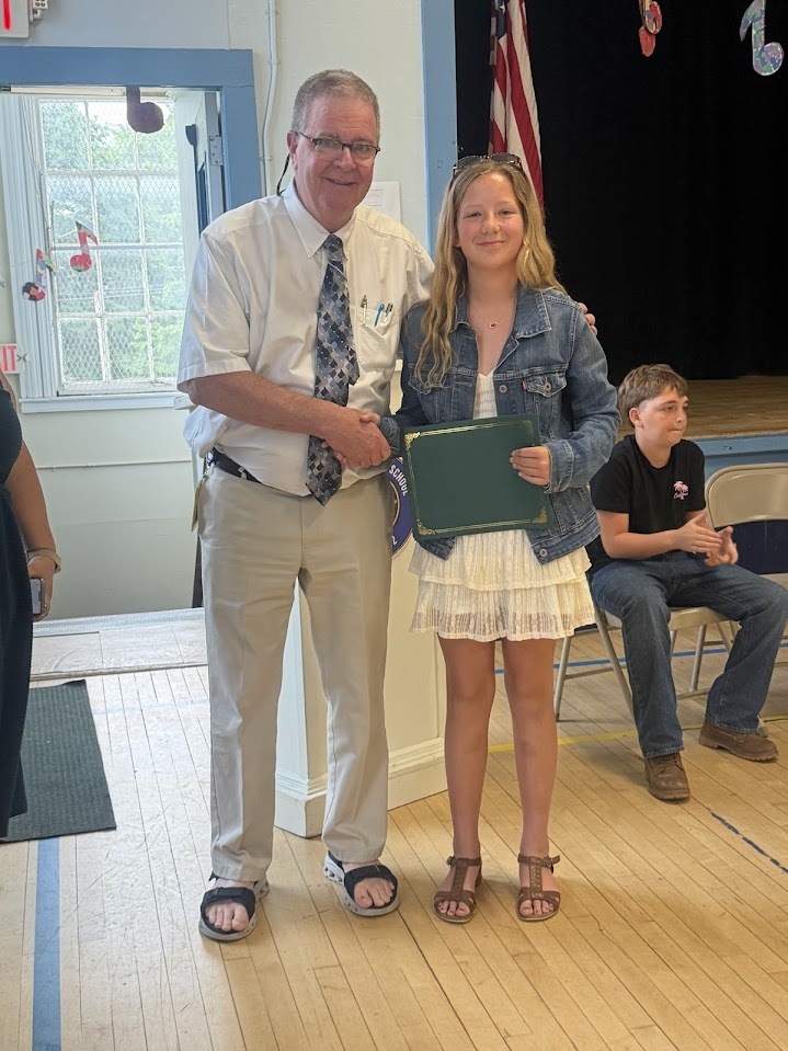 A sixth-grade student in a white dress and denim jacket smiles while receiving a certificate and congratulations from an administrator during a school recognition ceremony.