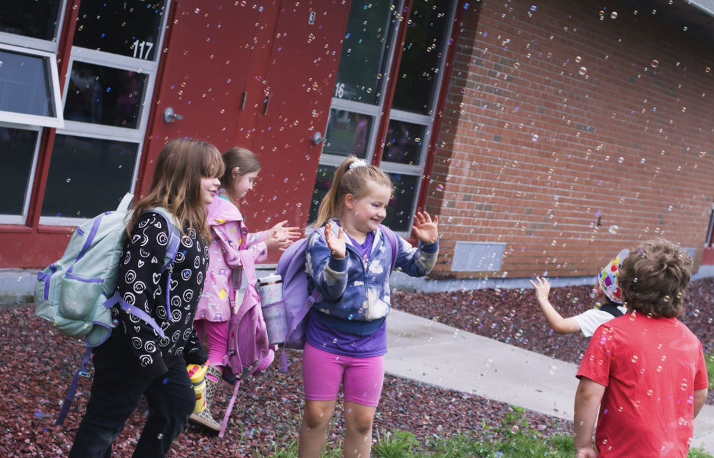 Young students wearing backpacks laugh and reach for bubbles floating outside the school building.