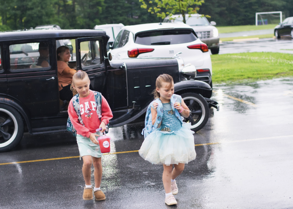Two young students walk across a wet parking lot after arriving in a vintage black car, one wearing a sparkly blue dress and the other carrying a backpack.