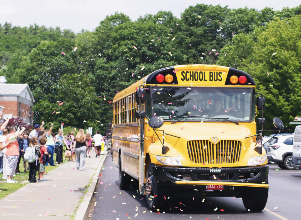 A school bus drives through a shower of confetti while students and staff cheer from the sidewalk during dismissal.