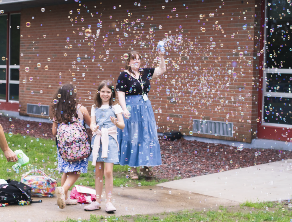 A staff member uses a bubble machine as students arrive at school, filling the air with hundreds of bubbles.