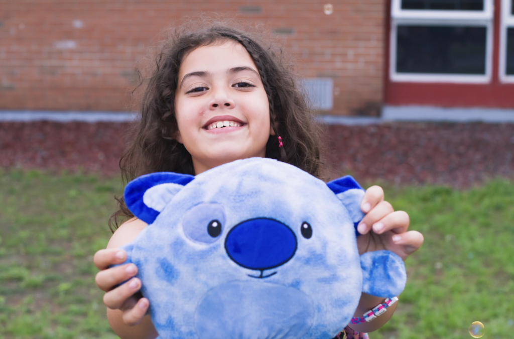 A smiling student proudly holds a blue stuffed animal while standing outside the school.