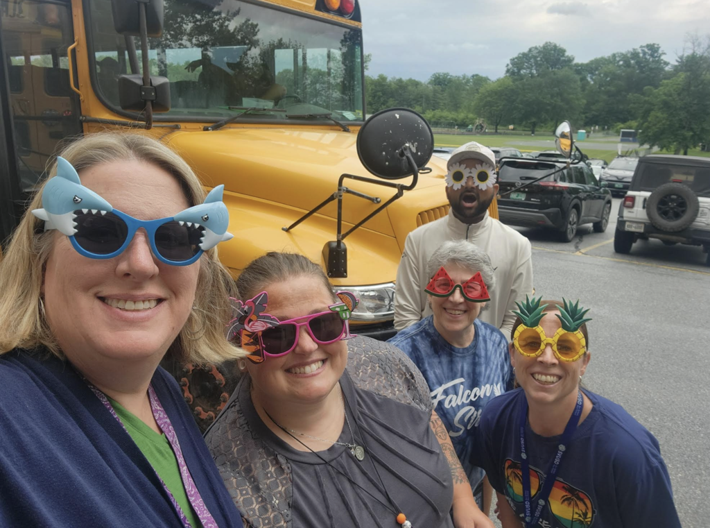 Five Fair Haven Grade School staff members pose in front of a school bus wearing playful novelty sunglasses shaped like sharks, pineapples, watermelons, flowers, and flamingos as they celebrate the last day of school and wish students a happy summer.
