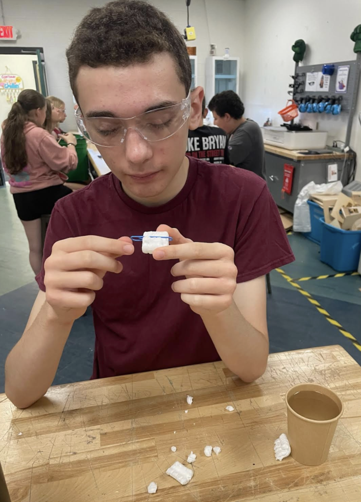 A student wearing safety glasses examines a small handmade device constructed from packing foam and craft materials during a classroom STEM activity.