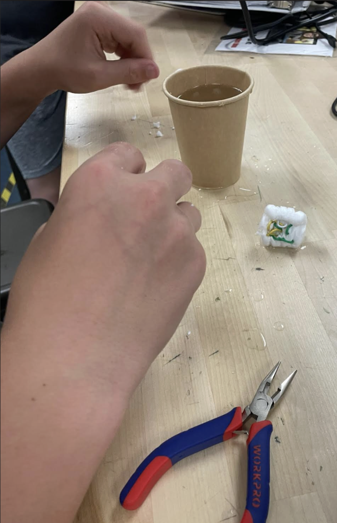 A close-up of a student's hands working on a small engineering project beside a paper cup of water, with needle-nose pliers and building materials spread across a workshop table.