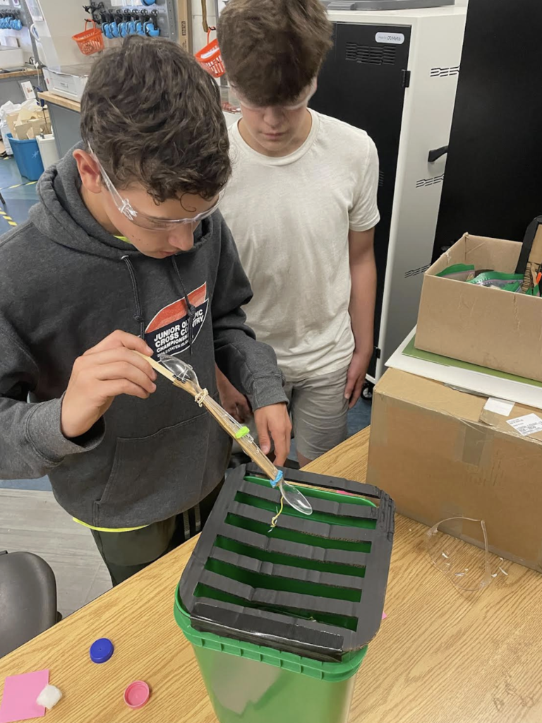 A student wearing safety glasses uses a handmade tool to lower an object through the slots of a green container while another student watches closely during a classroom engineering challenge.