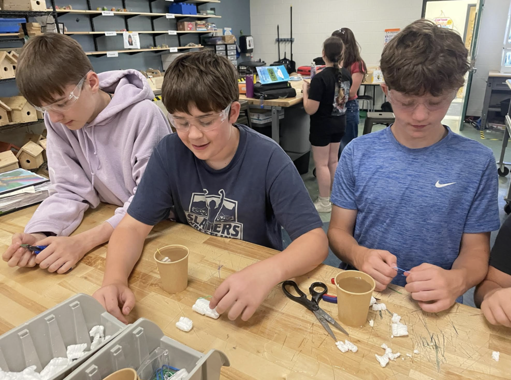 Three students wearing safety glasses work together at a workshop table, building and testing small devices using cups, packing materials, and craft supplies in a technology classroom.