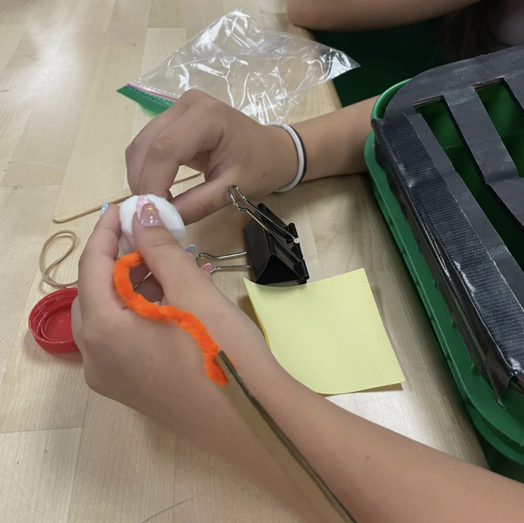 A close-up shows a student using cotton, a pipe cleaner, binder clip, and other materials to build a small prototype next to a green container with a slotted lid.