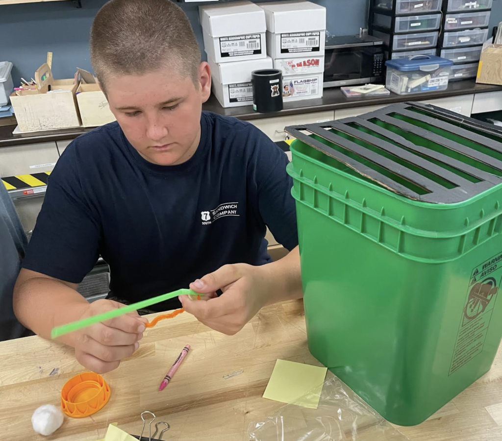 A student concentrates on attaching materials to a green straw while working on an engineering design challenge at a classroom workbench, with a green container and building supplies nearby.