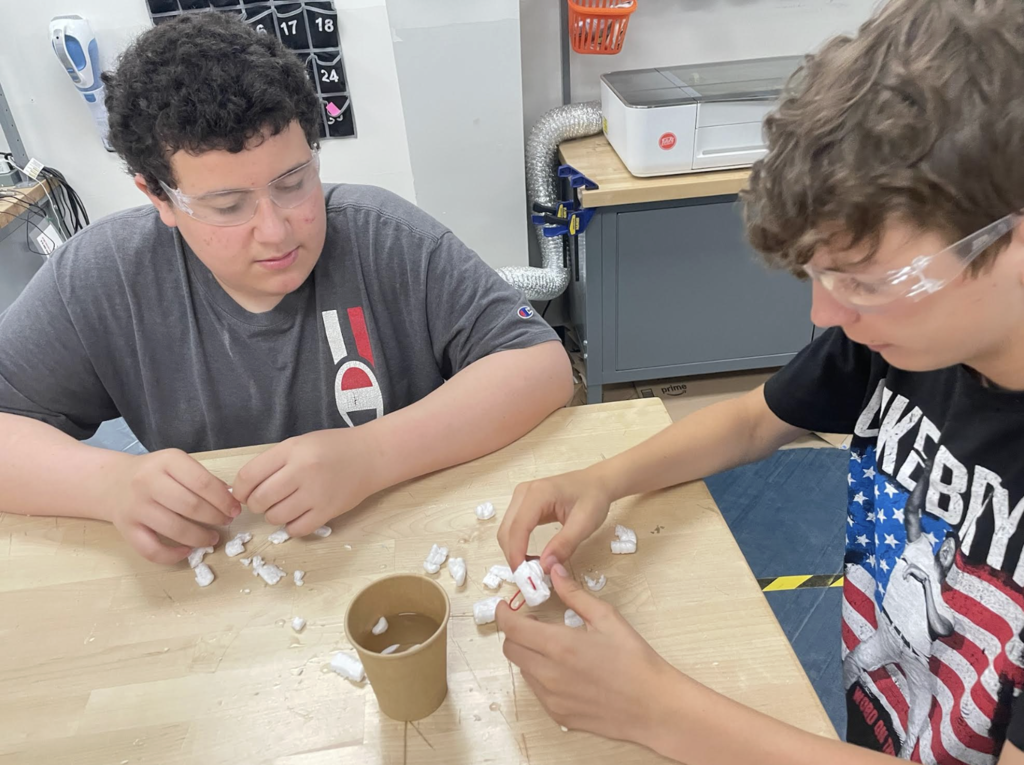 Two students wearing safety glasses carefully assemble a small project made from packing foam and other materials while seated at a worktable in a technology lab.