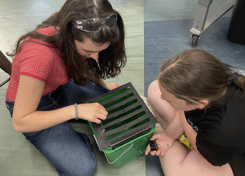 Two students wearing safety glasses sit on the floor working together to assemble a device on top of a green plastic container with a slotted black lid during a STEM or engineering activity.
