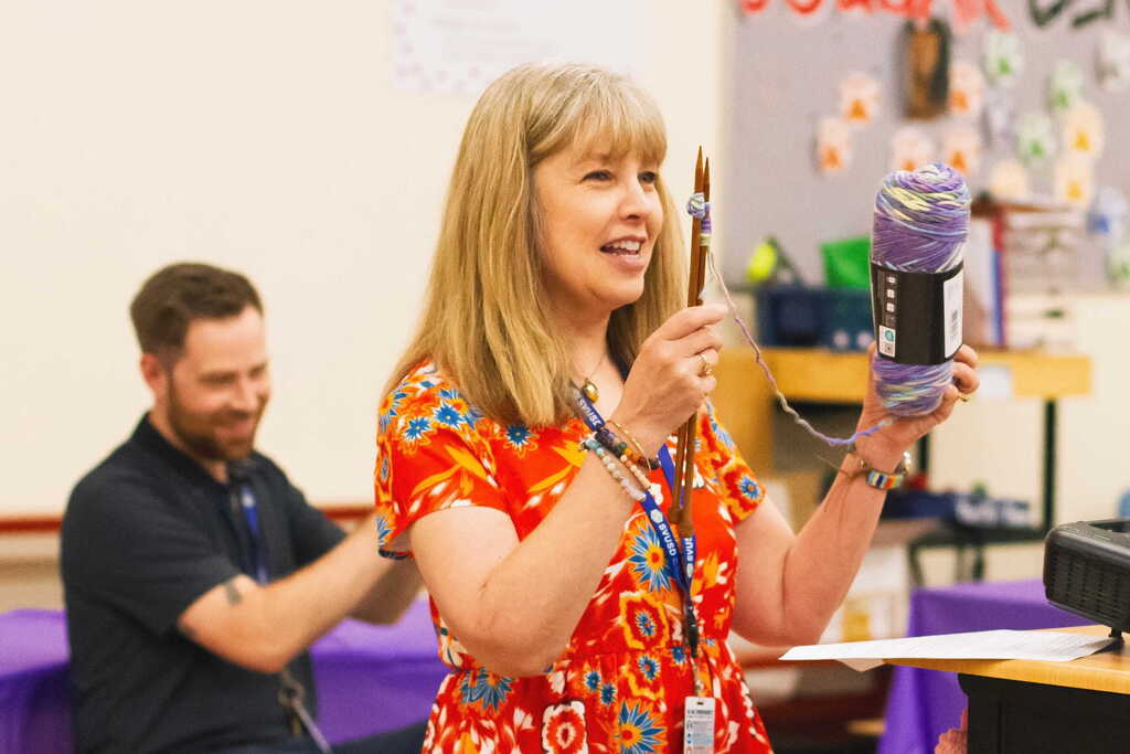 A colleague presents a skein of yarn and knitting needles while speaking to the group during the knitting-themed retirement celebration for Tina Rampone.