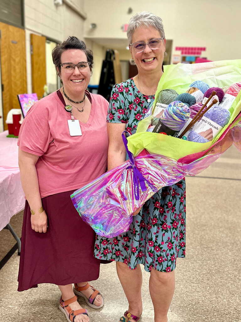 A colleague stands beside Tina Rampone, smiling while presenting her with a bouquet made of colorful yarn and knitting supplies during her retirement celebration.