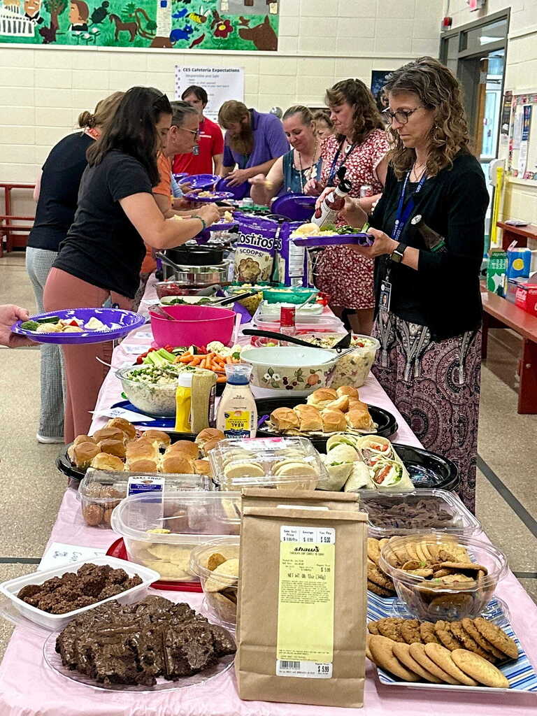 Faculty and staff gather around a long potluck buffet table filled with salads, sandwiches, desserts, and homemade dishes during Tina Rampone's retirement luncheon.