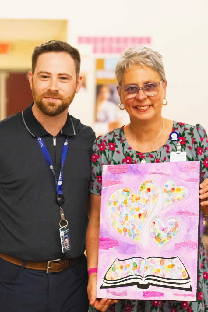The school principal stands beside retiring librarian Tina Rampone as she holds a colorful student-created artwork featuring hearts and an open book during her retirement celebration.