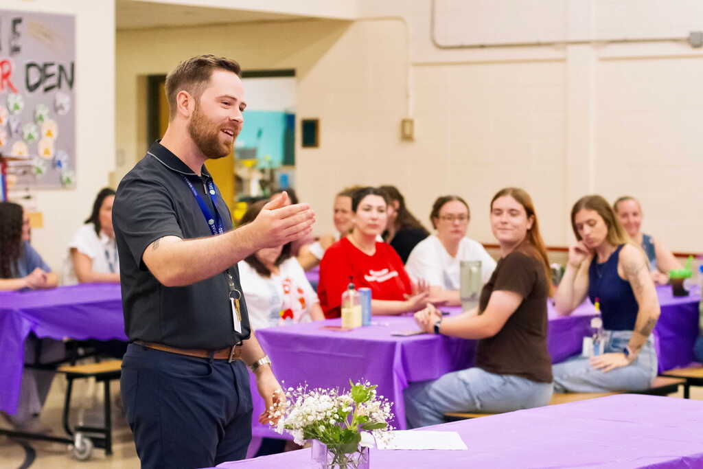 The school principal addresses faculty and staff gathered for Tina Rampone's retirement luncheon as attendees listen from tables decorated in purple.